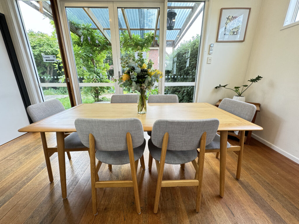 A bright and welcoming therapy group room with a large wooden table and comfortable gray chairs, a vase of yellow flowers adding warmth, situated in the Balanced: Mind Body Life clinic in Hobart