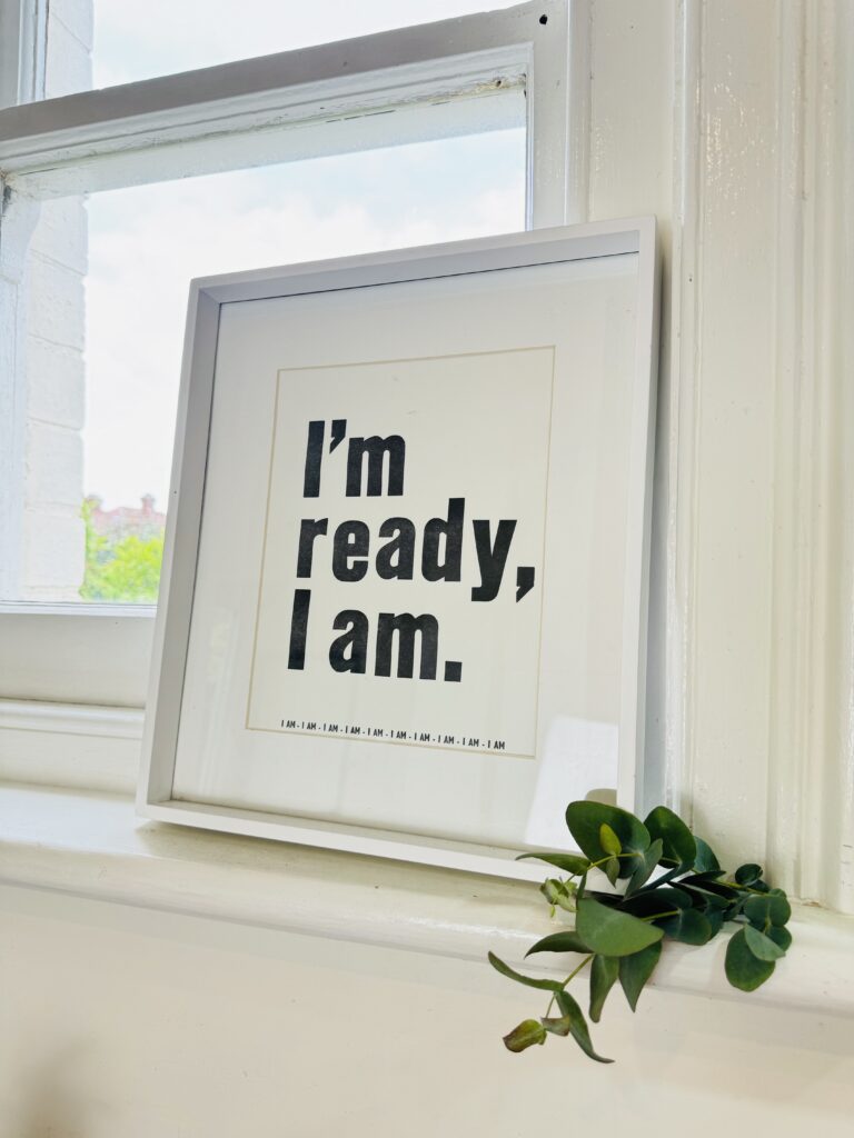 A framed print sits on a white windowsill, displaying the motivational phrase 'I'm ready, I am.' in bold black letters on a white background. Beneath the frame, a sprig of greenery adds a touch of natural color to the serene setting