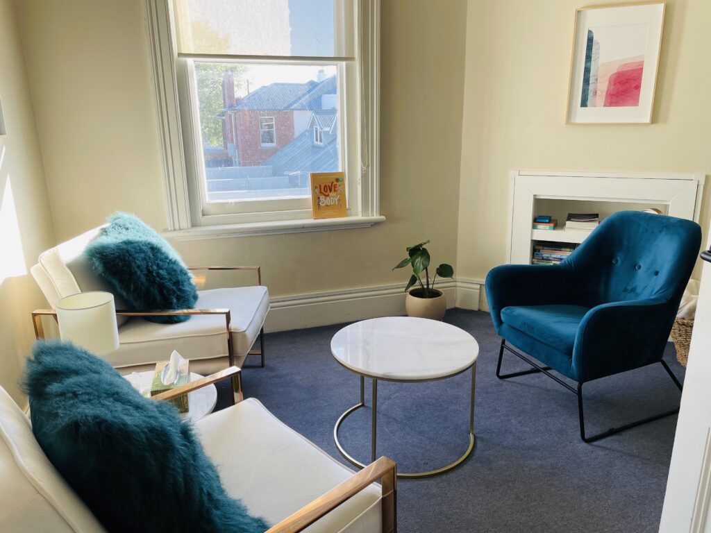 An intimate and serene consultation room at Balanced: Mind Body Life in Hobart, with a plush blue chair, white couches with teal pillows, a white marble coffee table, promoting a calming environment for clients