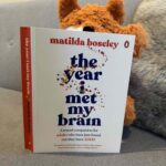A picture of the book 'The Year I Met My Brain' held up by a weighted plushie fox